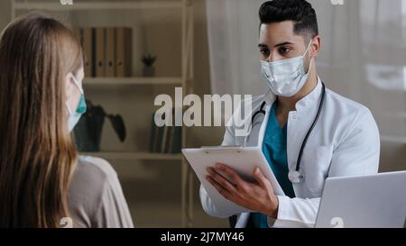 Arabischer männlicher Arzt Arzt in medizinischer Gesichtsmaske Notizen auf Zwischenablage Beratung weibliche Patientin erklären medizinische Ergebnisse geben Rezept Stockfoto