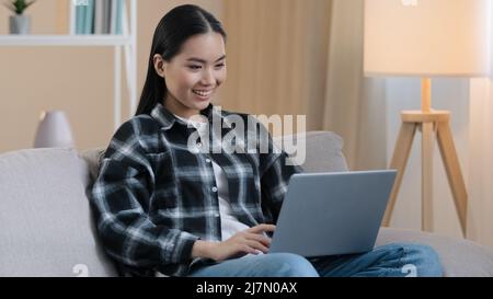 Lächelnde asiatische Frau glücklich Student Mädchen Freiberufler sitzt auf der Couch mit Laptop Eingabe Nachricht E-Mail Dame chatten auf Computer Surfen Internet-Medien Stockfoto