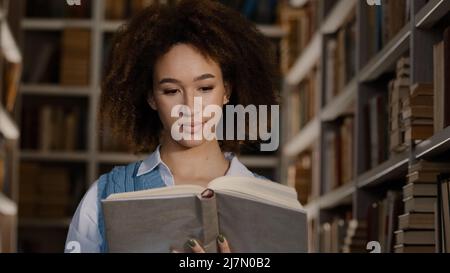 Junge afroamerikanische Mädchen Student steht in der öffentlichen Universitätsbibliothek interessiert an liest Buch interessante Geschichte sorgfältig Blick auf Lehrbuch Stockfoto