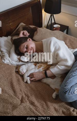 Junge Frau mit niedlichen Ingwer Katze mit grünen Augen. Weiblich umarmt ihre niedliche kurze Haare Kitty im Schlafzimmer. Platz kopieren, Nahaufnahme. Entzückendes Haustier Stockfoto