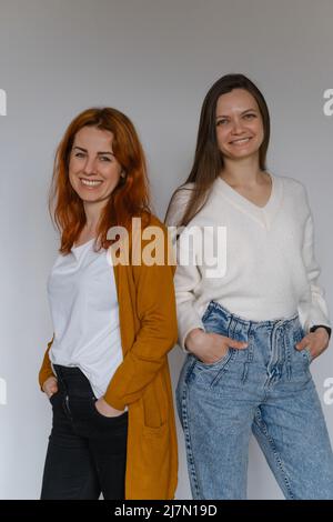 Glückliche zwei Frauen im Alter von 20s 40s über einer weißen Wand. Mitarbeiter in legerer Kleidung, die auf die Kamera schauen. Frau mit langen Haaren, fröhliche Freiberuflerin Stockfoto