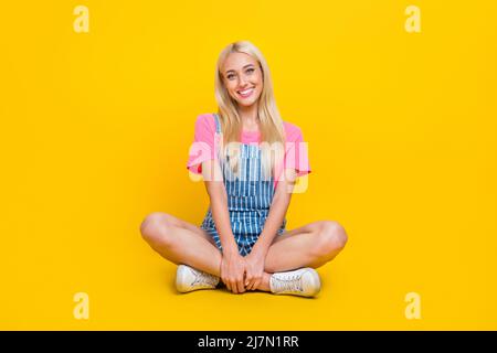 Foto in voller Größe von schönen zufriedenen Mädchen sitzen Boden gekreuzte Beine toothy Lächeln isoliert auf gelbem Hintergrund Stockfoto