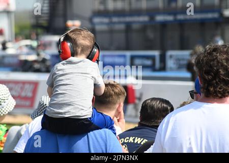 Ein Kind saß bei einem Event mit roten Kopfhörern auf den Schultern eines mannes, Ohrenschützer auf dem Kopf in einer Menschenmenge von hinten Stockfoto