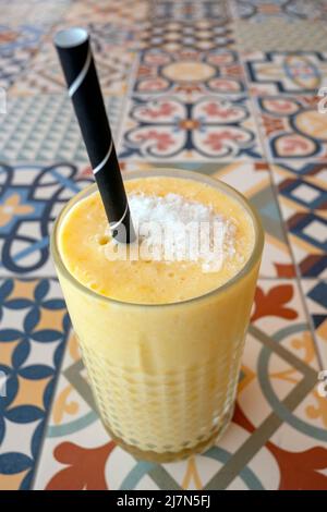 Großes Glas frisch zubereiteter Ananas und Mango-Smoothie mit Kokosflocken auf der Oberseite und einem Papierstroh für einen einfachen, gesunden Fruchtdrink Stockfoto