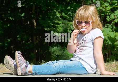 Süßes Kind, das draußen auf einer Parkbank sitzt und einen Lutscher saugt Stockfoto