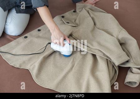 Frau verwendet eine Maschine, um Pellets und Spulen aus Kleidung und Stoff auf schwarzen Hosen zu entfernen. Ein modernes elektronisches Gerät zur Aktualisierung alter Dinge Stockfoto