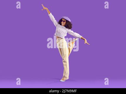 Happy Black Mädchen im lässigen Outfit stehen auf einem Fuß auf lila Studio Hintergrund Stockfoto