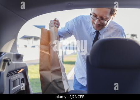 Mann, der auf dem Parkplatz eines Einkaufszentrums war. Junger kaukasischer lächelnder Mann, der Papiertüten mit Essen in den Kofferraum legte. Stockfoto