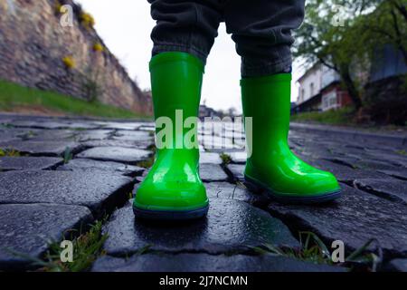 Ein kleines Kind in grünen Gummistiefeln steht auf einer asphaltierten Straße. Foto der Füße eines Kindes in Gummischuhen auf dem Bürgersteig nach Regen. Stockfoto