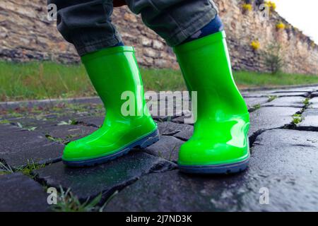 Ein kleines Kind in grünen Gummistiefeln steht auf einer asphaltierten Straße. Foto der Füße eines Kindes in Gummischuhen auf dem Bürgersteig nach Regen. Stockfoto