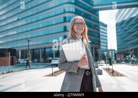 Porträt eines glücklichen jungen Büroassistenten, der aus der Ferne mit einem Laptop im Freien arbeitet, während er mit einer Brille vor einem Gebäude posiert Stockfoto