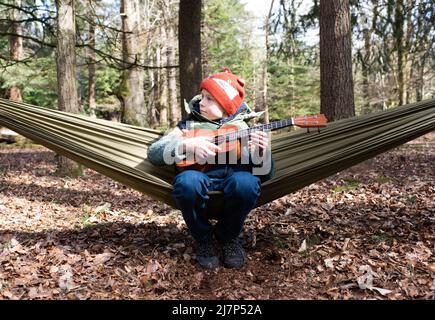 Junge saß in einer Hängematte im Wald und spielte die Ukelele entspannend Stockfoto