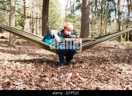 Junge spielt fröhlich Gitarre in einer Hängematte im Wald Stockfoto