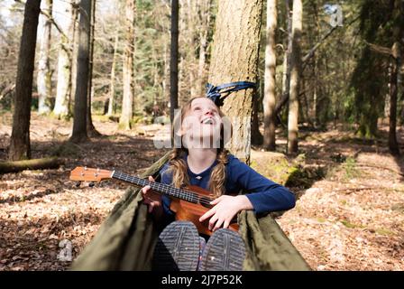 Das Kind saß in einer Hängematte und spielte die Ukulele im Wald Stockfoto