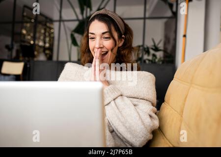 Aufgeregte Frau, die zu Hause auf dem gemütlichen Sofa auf den Laptop-Bildschirm schaut Stockfoto