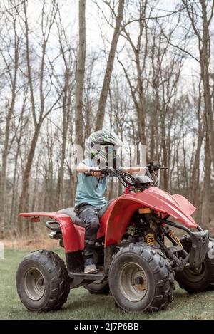 Junge auf einem roten Vierrad im Gras mit Wald im Hintergrund Stockfoto