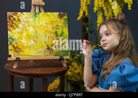 Ein Mädchen sitzt in der Nähe einer Mimose und malt ein Bild in einem blauen Kleid Mimosa Pinsel wenig niedlich, am Nachmittag Porträt hält in der Kindheit und Blume Stockfoto