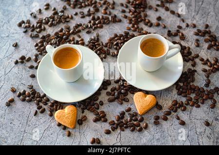 Zwei Tassen Kaffee auf Untertassen, umgeben von Kaffeebohnen und herzförmigen Keksen. Das Arrangement schafft eine warme und einladende Atmosphäre. Stockfoto