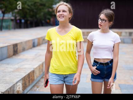 Porträt von zwei positiven Mädchen, die entlang der Straße der Sommerstadt gehen Stockfoto