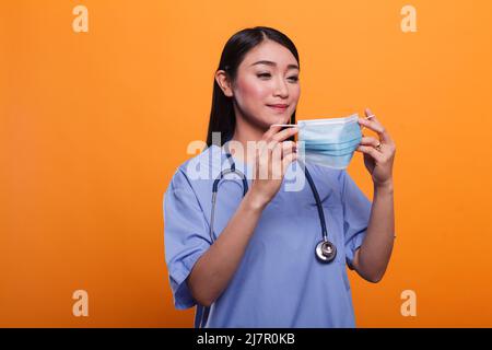 Healthcare Klinik asiatische Krankenschwester in blauer Uniform, trägt Stethoskop setzen auf Gesichtsmask gegen covid. Pflegekraft des Krankenhauses, die beim Tragen eines medizinischen Instruments eine Virusschutzmaske aufsetzt. Stockfoto