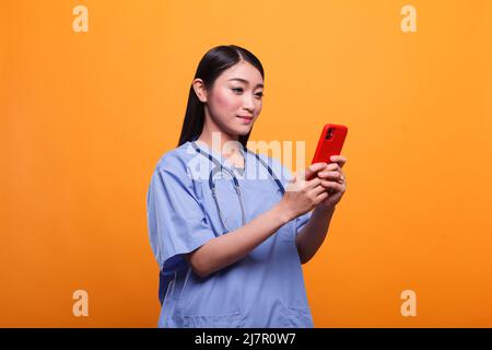 Selbstbewusste Krankenschwester im Gesundheitswesen Asiatische Krankenschwester trägt Stethoskop, während sie Smartphone zum Senden von Nachrichten verwendet. Gut aussehende Pflegekraft mit Handy-Smart-Gerät und medizinisches Instrument. Stockfoto