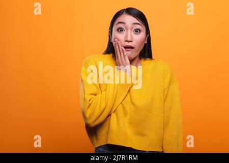 Schockiert beunruhigte asiatische Frau trägt gelben lebhaften Pullover, während die Hand auf das Gesicht. Unbequem jungen erwachsenen Mädchen mit Schock Ausdruck, trägt lebendige trendige Kleidung, während auf orangefarbenem Hintergrund. Stockfoto