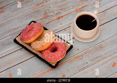 Drei Donuts in weißer und rosafarbener Glasur auf einem schwarzen Teller und eine Tasse schwarzen Kaffee auf einem Holztisch. Draufsicht, flach liegend. Stockfoto