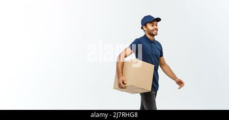 Lieferkonzept - Portrait eines glücklichen afroamerikanischen Lieferers in blauem Stoff, der zu Fuß ein Paket an den Kunden schickt. Isoliert im Gray Studio Stockfoto