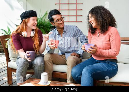 Eine Gruppe von multirassischen Freunden mit Tassen Kaffee plaudert und schaut sich gegenseitig an, während sie auf einem bequemen Sofa auf der hellen Terrasse sitzen Stockfoto