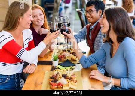 Eine Gruppe fröhlicher multirassischer Freunde, die sich gegenseitig angucken und mit Wein in die Gläser klirren, während sie ein festliches Ereignis am Tisch mit Essen auf der Terra feiern Stockfoto