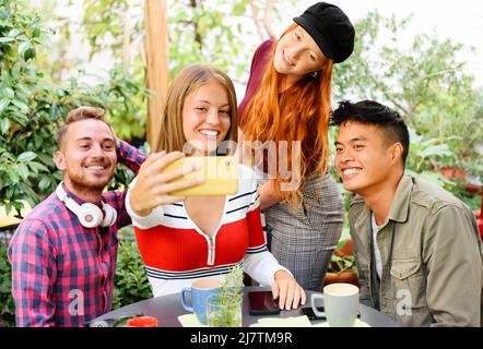Lächelnde, vielfältige Freunde in legerer Kleidung treffen sich mit Tassen Getränken um den Tisch und porträtieren sich selbst auf dem Smartphone auf der Terrasse des Cafés. Lebst Stockfoto