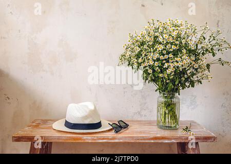 Strohhut, Sonnenbrille und ein Strauß Gänseblümchen auf einer Holzbank Stockfoto