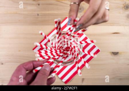 Nahaufnahme einer Menge von Trinkhalmen in einer Glasschale auf einem Holztisch - Hände nehmen die Trinkhalme - Studioaufnahme Stockfoto