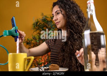 Die schöne Frau mit lockigen Haaren plaudert mit dem Smartphone, das an einem Telefonhalter hängt, beim Frühstück Stockfoto