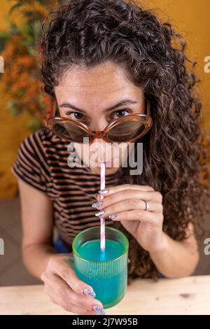 Hübsche Frau mit Vintage-Sonnenbrille sitzt auf einem Tisch und trinkt blauen Cocktail durch einen Stroh auf farbigem Hintergrund Stockfoto