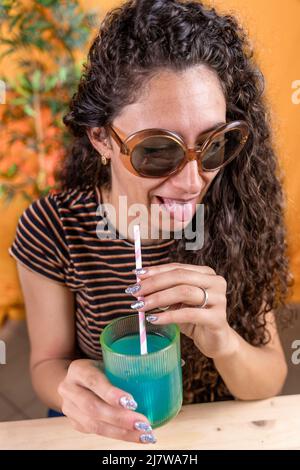 Hübsche Frau mit Vintage-Sonnenbrille sitzt auf einem Tisch und trinkt blauen Cocktail durch einen Stroh auf farbigem Hintergrund Stockfoto
