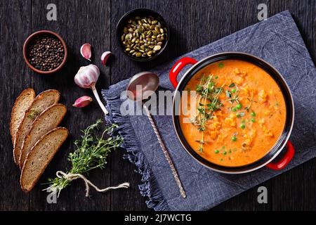 Dicke Süsskartoffelsuppe mit Gemüse in rotem Topf auf dunklem Holztisch mit Kräutern, Pfefferkörnern, Kürbiskernen, Roggenbrotscheiben und frischem Thymian, Stockfoto