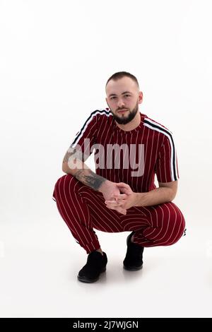 Dunkelhaarige isolierte bärtige gutaussehende fröhlich attraktiv selbstbewussten Mann sitzt in Hocke Pose auf dem Boden in roter Uniform Stockfoto