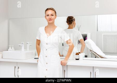 Eine junge lächelnde Kosmetologin, die in einem medizinischen Büro neben dem Tisch posiert. Das Konzept der professionellen Kosmetologie. Stockfoto