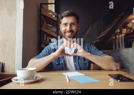 Porträt eines glücklichen jungen, gutaussehenden Mannes, der im Café mit Händen einen Herzensanschlag zeigt. Liebe Demonstration. Stockfoto