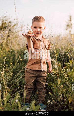 Ein kleiner Junge steht in der Natur im Gras und hält eine Schnecke in der Hand Stockfoto