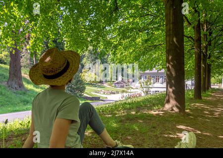 Frau mit braunem Haar, grauem T-Shirt, Jeans und Strohhut sitzt auf einer Wiese unter dem Schatten eines Baumes und blickt auf den Staatspark Fürstenlager Auer Stockfoto