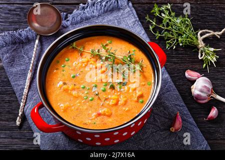 Gebratene, dicke Yamssuppe mit Gemüse in rotem Topf auf einem dunklen Holztisch mit Suppenkelle, horizontale Ansicht von oben, Nahaufnahme Stockfoto