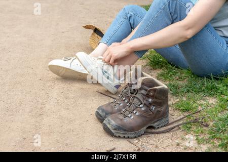 Frau, die Jeans trägt, ihre Schuhe wechselt, ihre normalen Schuhe auszieht und ihre braunen Wanderschuhe anzieht, die neben ihr stehen Stockfoto