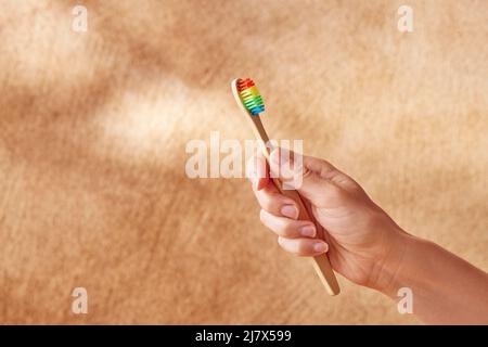 Crop unkenntlich weiblich zeigt umweltfreundliche Bambus-Zahnbürste mit bunten Borsten vor verschwommenem beigen Hintergrund Stockfoto