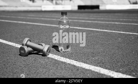 Langhanteln und Flasche auf der Laufstrecke. Sportgerät und -Ausrüstung. Kurzhanteln auf der Rennstrecke im Stadion. Hydratation nach Fitness. Gesunder, ausgewogener Lebensstil Stockfoto