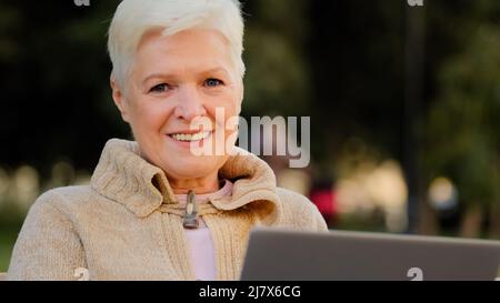 Lächelnde ältere Frau im Rentenalter, die am Laptop arbeitet, um online Nachrichten zu lesen, ältere Dame, die am pc im Freien eine SMS schreibt. Glücklich gealterte Frau mit einem Stockfoto