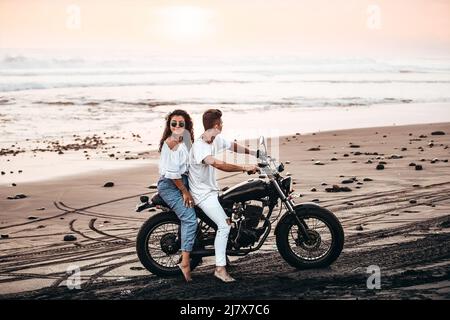 Männliches und weibliches Paar in weißem Outfit, das während des Sonnenuntergangs auf einem Motorrad an einem schwarzen Sandstrand in Bali Indonesien auf einem schwarzen Vintage-Motorrad sitzt Stockfoto