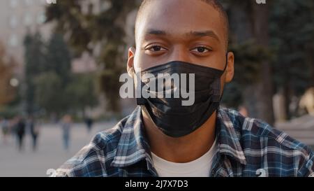 Nahaufnahme männlich Porträt afroamerikanischen Mann krank Kerl in medizinische Maske sitzt im Freien in der Stadt Straße in der Kamera in Quarantäne covid 19 Stockfoto
