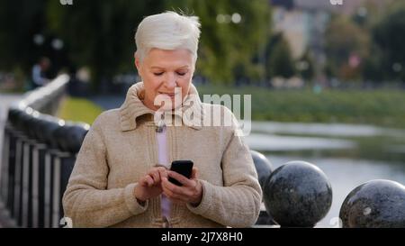 Lächelnd reifen 60s Frau SMS-Nachrichten auf moderne Smartphone-Gadget, glücklich Senior 70s Großmutter ruhen im Freien im Internet auf Handy surfen Stockfoto